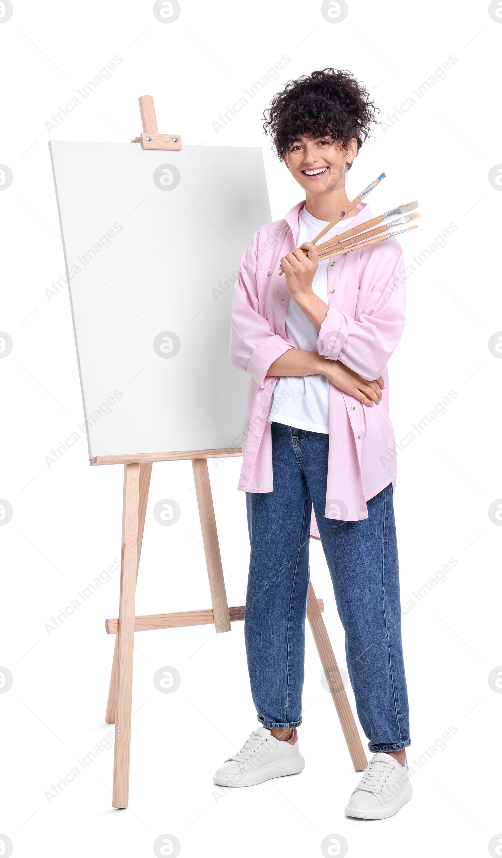 Photo of Young woman holding brushes near easel with canvas against white background