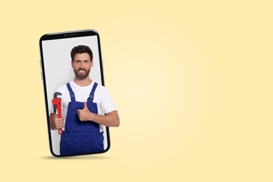 Image of Plumber looking out of smartphone and showing thumbs up on light yellow background. Space for text