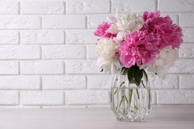 Beautiful peonies in glass vase on white table near brick wall. Space for text
