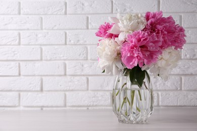 Photo of Beautiful peonies in glass vase on white table near brick wall. Space for text