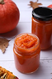 Jars of delicious pumpkin jam, fresh pumpkin and dry leaves on white wooden table, closeup