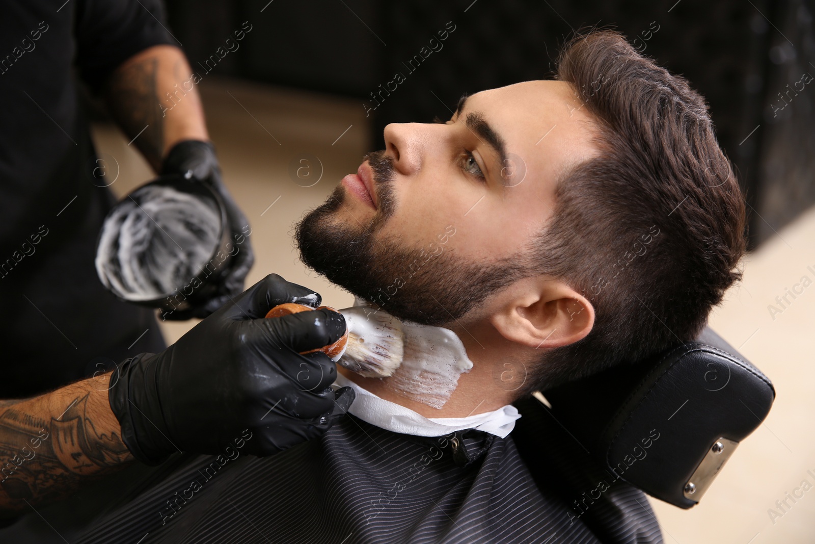 Photo of Professional hairdresser applying shaving foam onto client's skin in barbershop