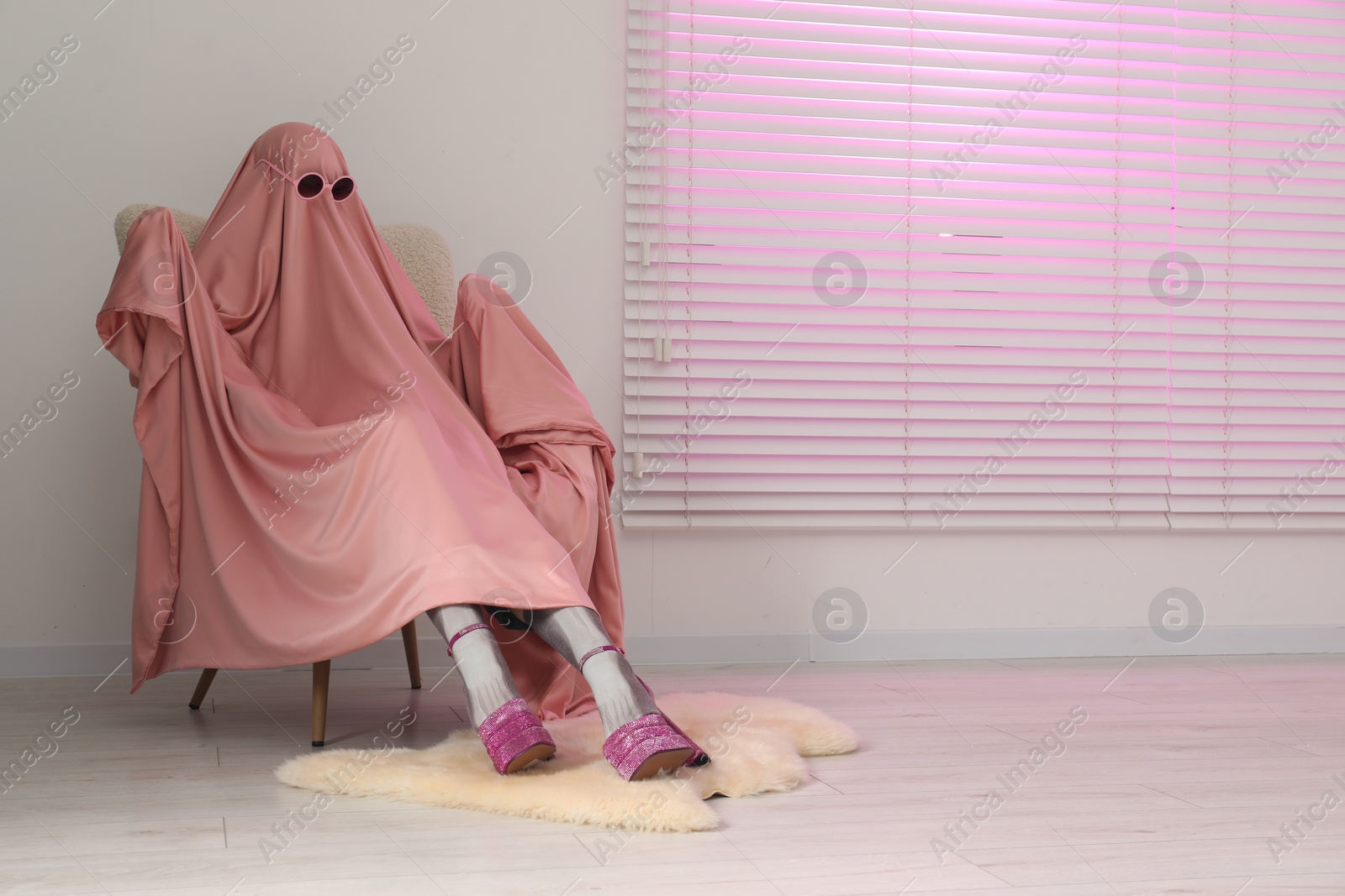 Photo of Glamorous ghost. Woman in pink sheet and high heel shoes on armchair indoors, space for text