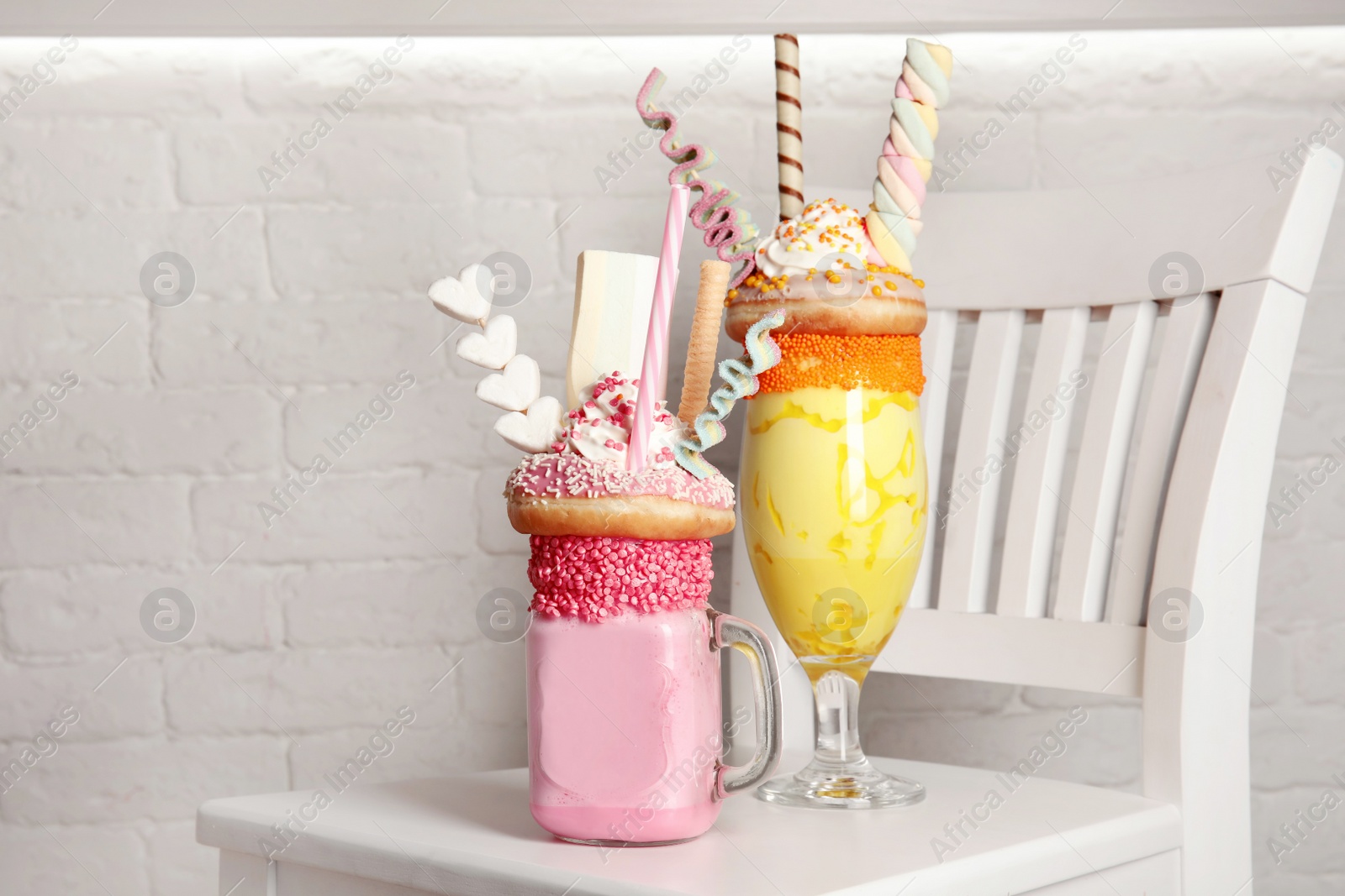 Photo of Tasty milk shakes with sweets in glassware on chair near brick wall