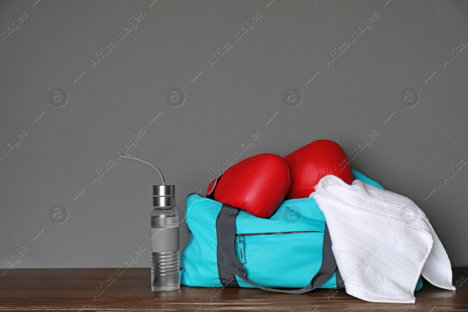 Photo of Sports bag, boxing gloves, towel and bottle of water on wooden table against gray background