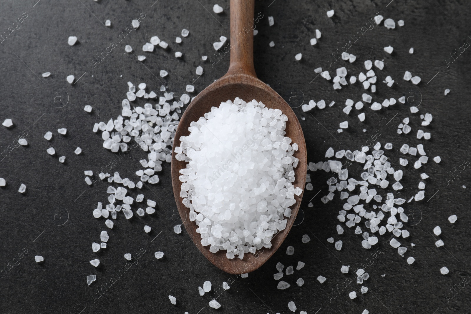 Photo of Natural salt and wooden spoon on black table, top view