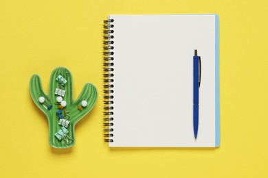 Ballpoint pen, notebook and different clips on yellow background, flat lay