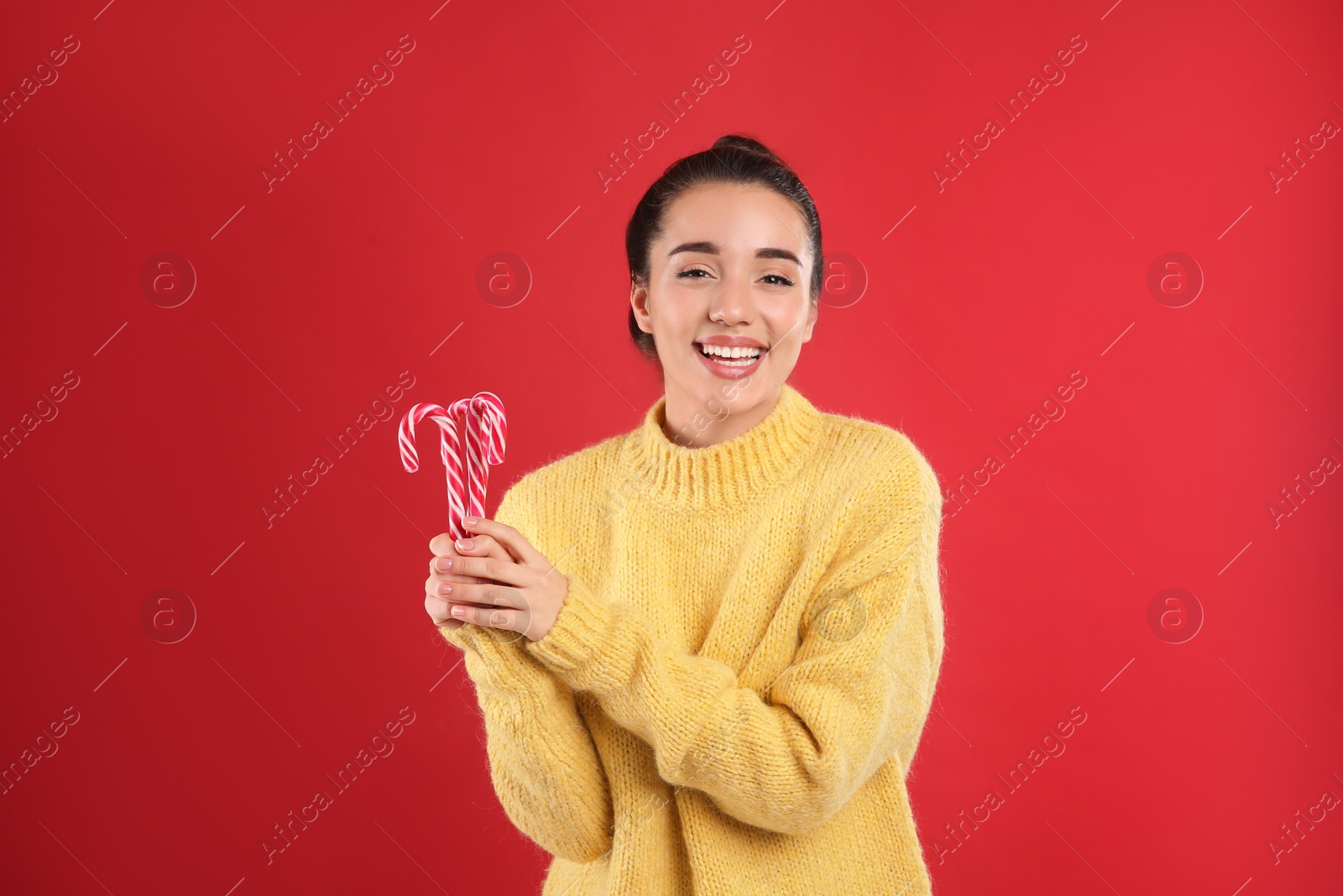Photo of Young woman in yellow sweater holding candy canes on red background. Celebrating Christmas
