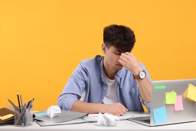 Tired young man working at white table on orange background. Deadline concept