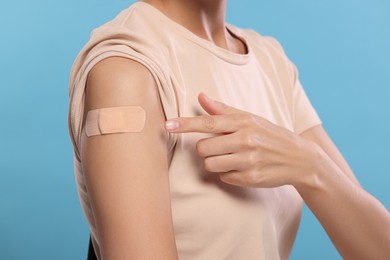 Woman pointing at sticking plaster after vaccination on her arm against light blue background, closeup