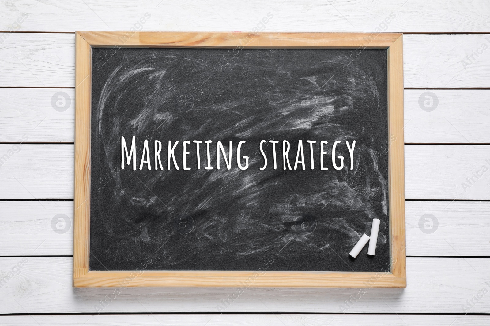 Image of Blackboard with pieces of chalk and phrase MARKETING STRATEGY on white wooden background, top view