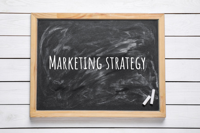 Image of Blackboard with pieces of chalk and phrase MARKETING STRATEGY on white wooden background, top view