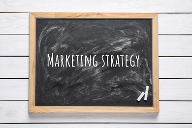 Blackboard with pieces of chalk and phrase MARKETING STRATEGY on white wooden background, top view