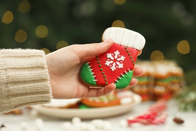 Photo of Woman with decorated cookie at table against blurred Christmas lights, closeup