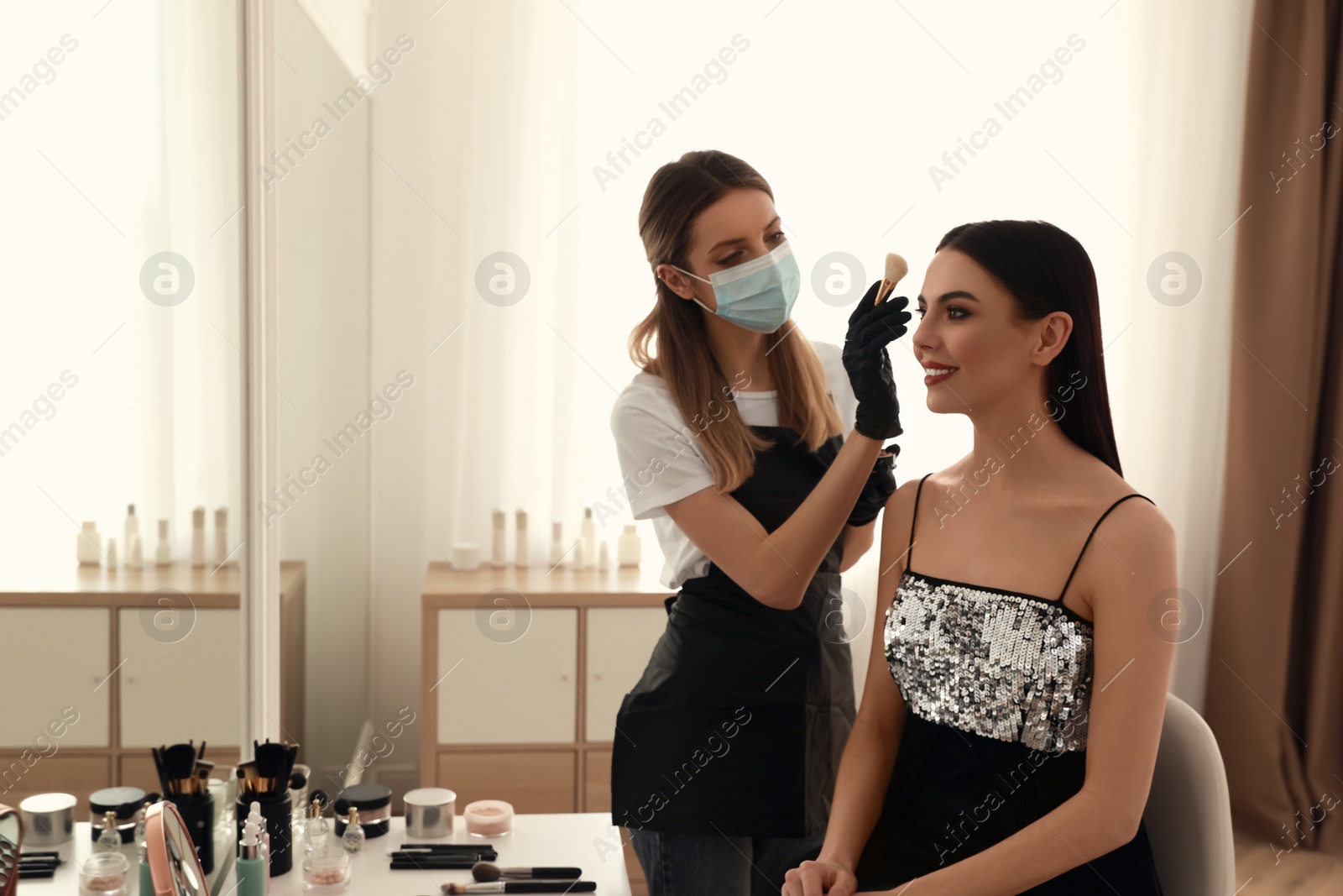 Photo of Makeup artist in protective mask and gloves working with woman indoors. Preventive measures during COVID-19 pandemic