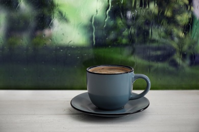 Photo of Cup of hot drink on wooden windowsill against glass with rain drops, space for text