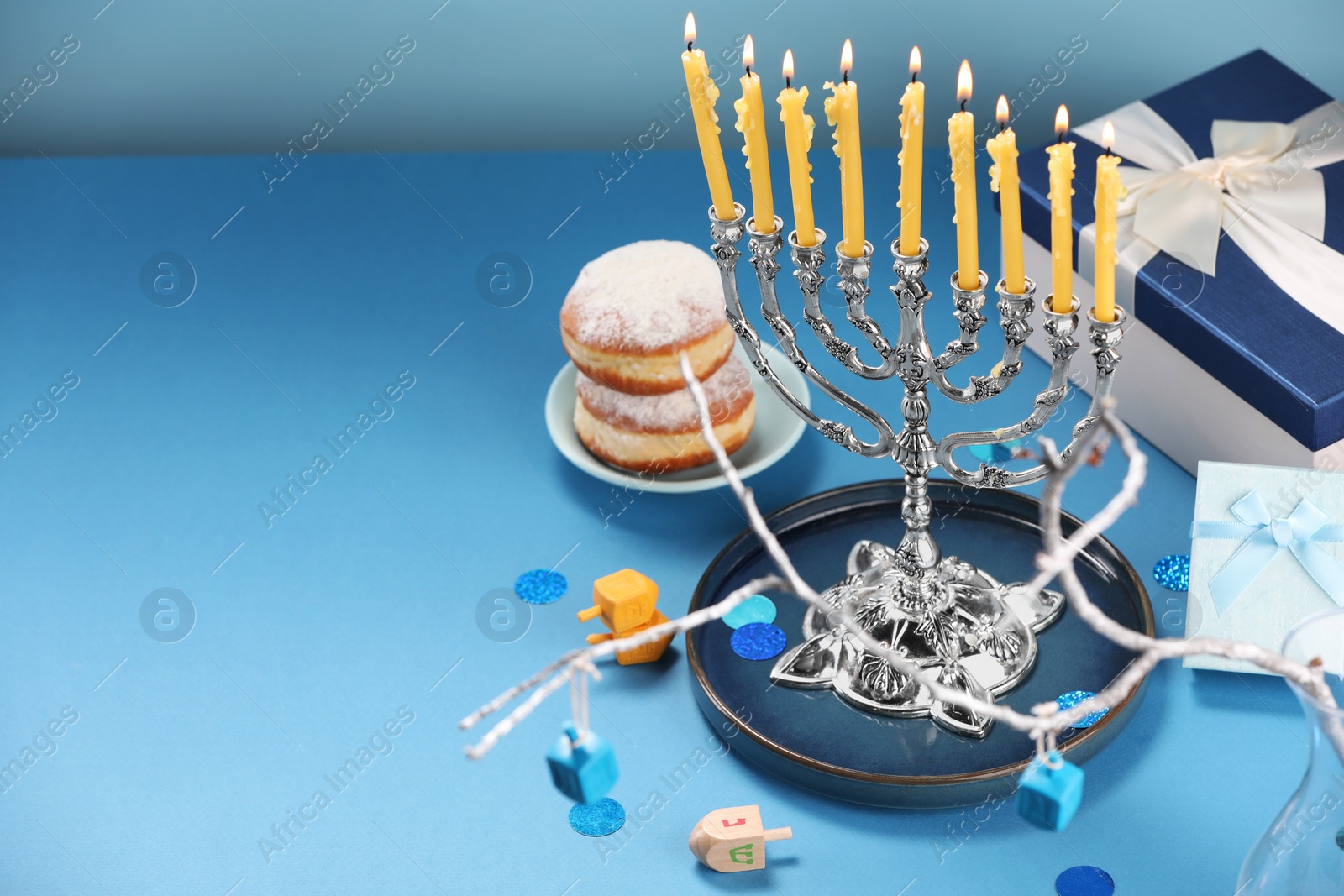 Photo of Hanukkah celebration. Menorah with burning candles, dreidels, donuts and gift boxes on light blue table. Space for text