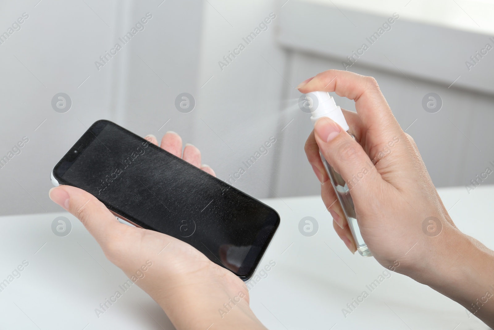 Photo of Woman spraying antiseptic onto smartphone at white table, closeup