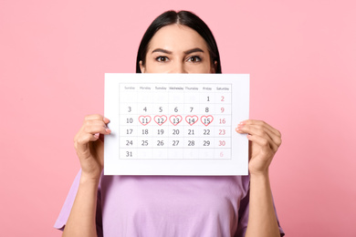 Photo of Young woman holding calendar with marked menstrual cycle days on pink background