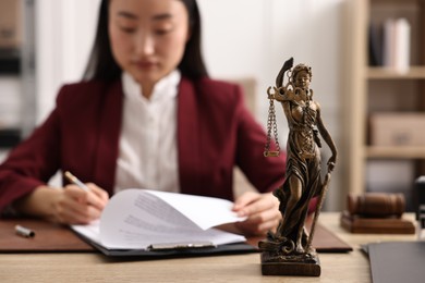 Photo of Notary signing document at table in office, focus on statue of Lady Justice