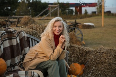 Beautiful woman sitting on wooden bench near pumpkins and hay bales outdoors. Autumn season