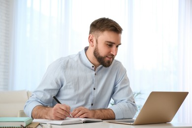 Photo of Young man taking notes during online webinar at table indoors