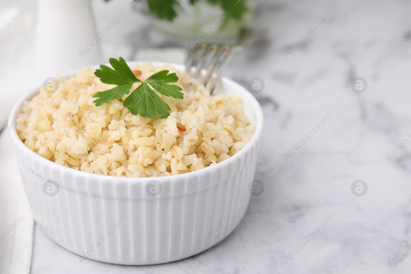 Photo of Cooked bulgur with parsley in bowl on white marble table, closeup. Space for text