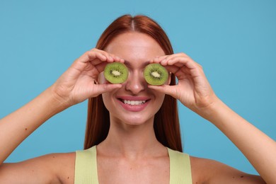 Photo of Happy woman covering eyes with halves of fresh kiwi on light blue background
