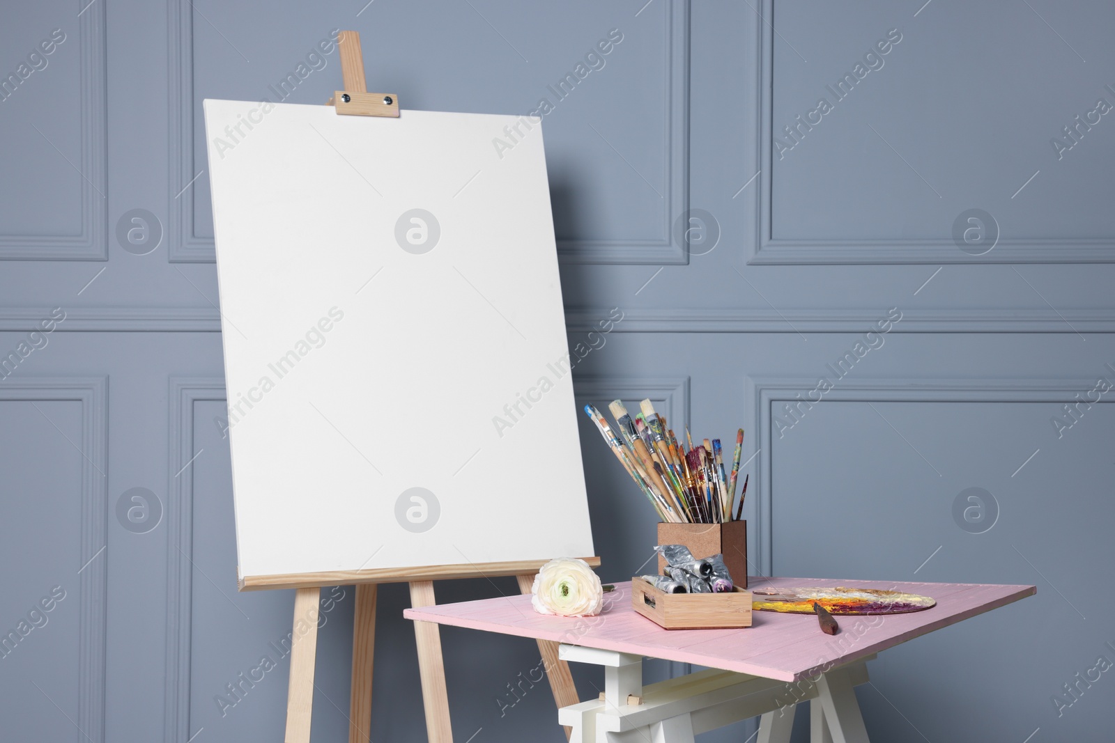 Photo of Easel with blank canvas and different art supplies on wooden table near grey wall