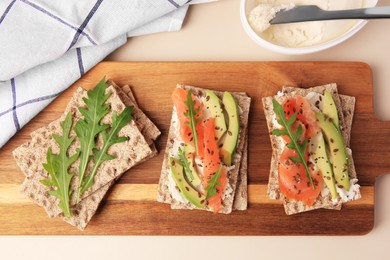 Fresh crunchy crispbreads with cream cheese, salmon, avocado and arugula on beige table, flat lay