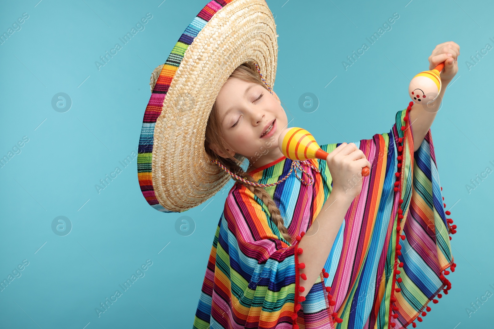 Photo of Cute girl in Mexican sombrero hat and poncho singing with maracas on light blue background