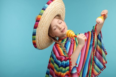 Photo of Cute girl in Mexican sombrero hat and poncho singing with maracas on light blue background