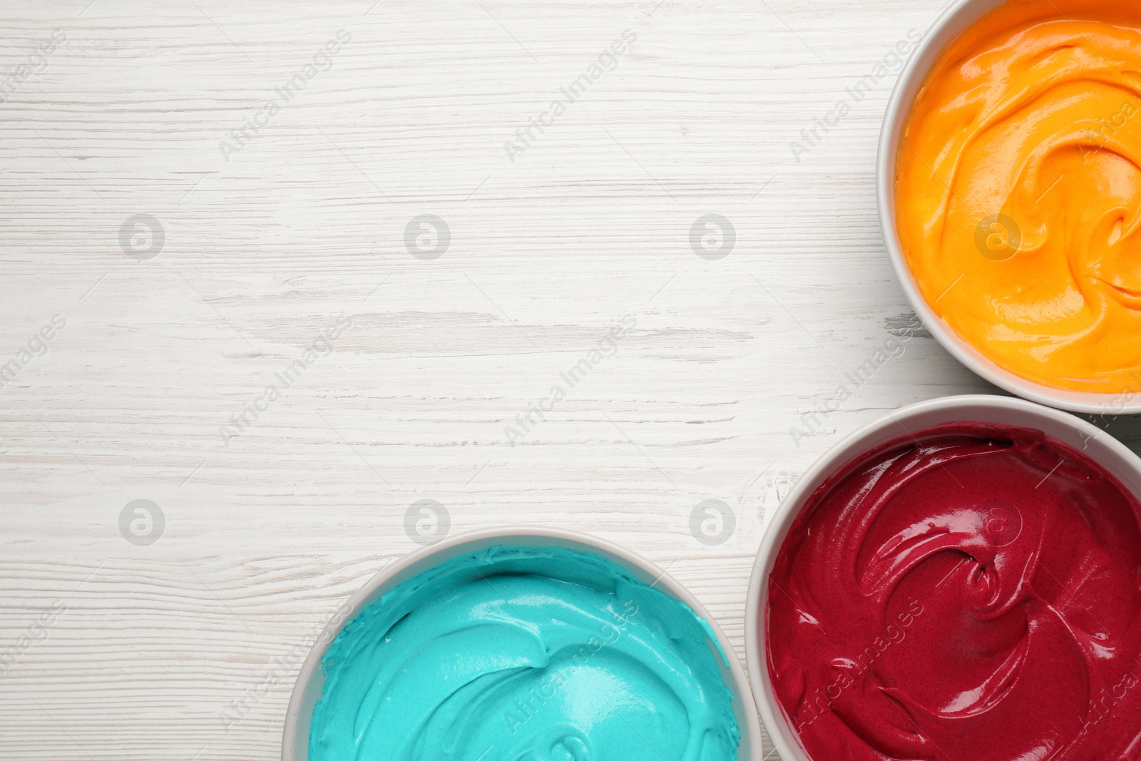 Photo of Bowls of different cream with food coloring on white wooden table, flat lay. Space for text