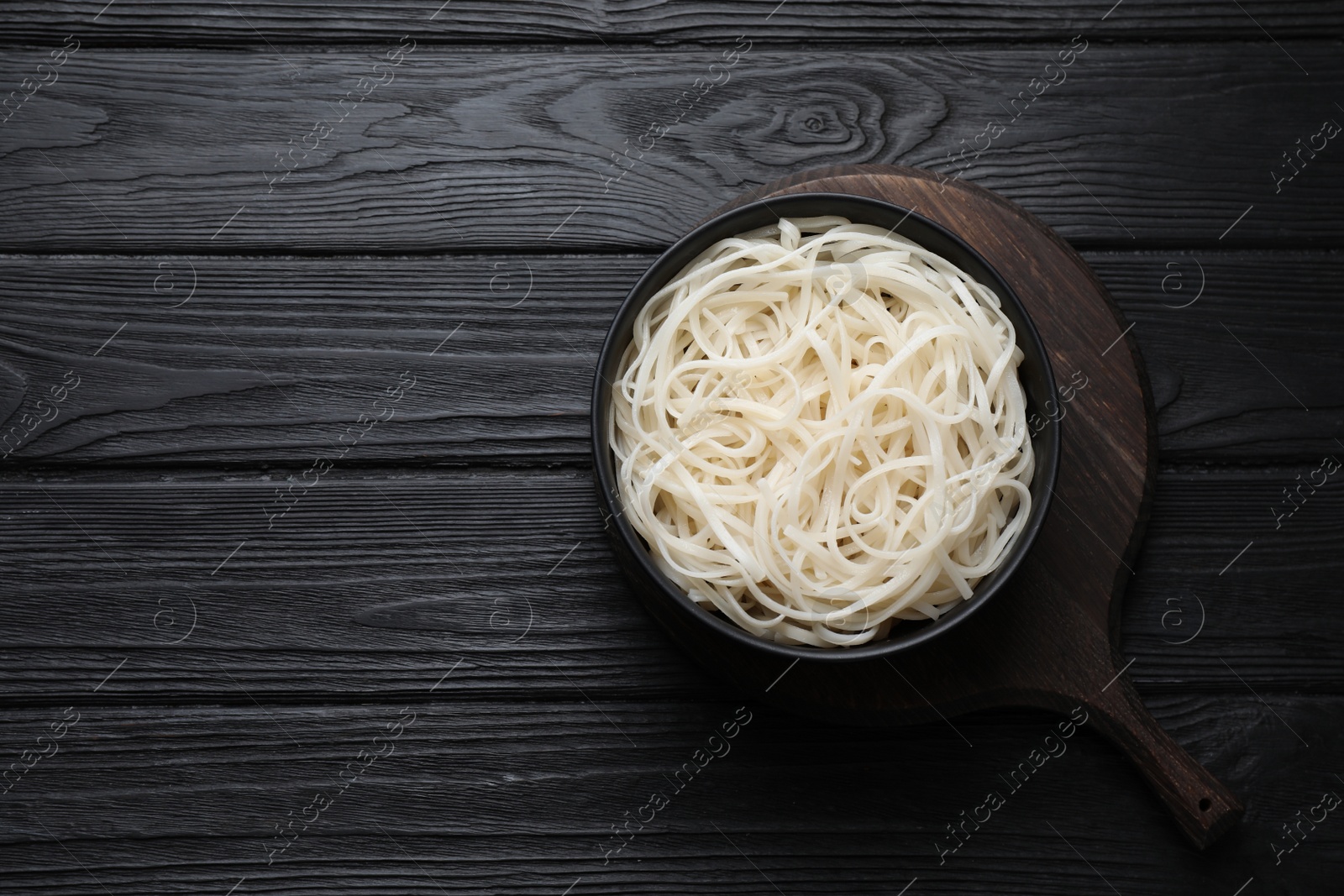 Photo of Bowl of tasty cooked rice noodles on black wooden table, top view. Space for text