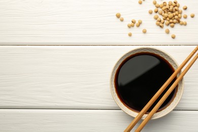 Tasty soy sauce in bowl, chopsticks and soybeans on white wooden table, top view. Space for text