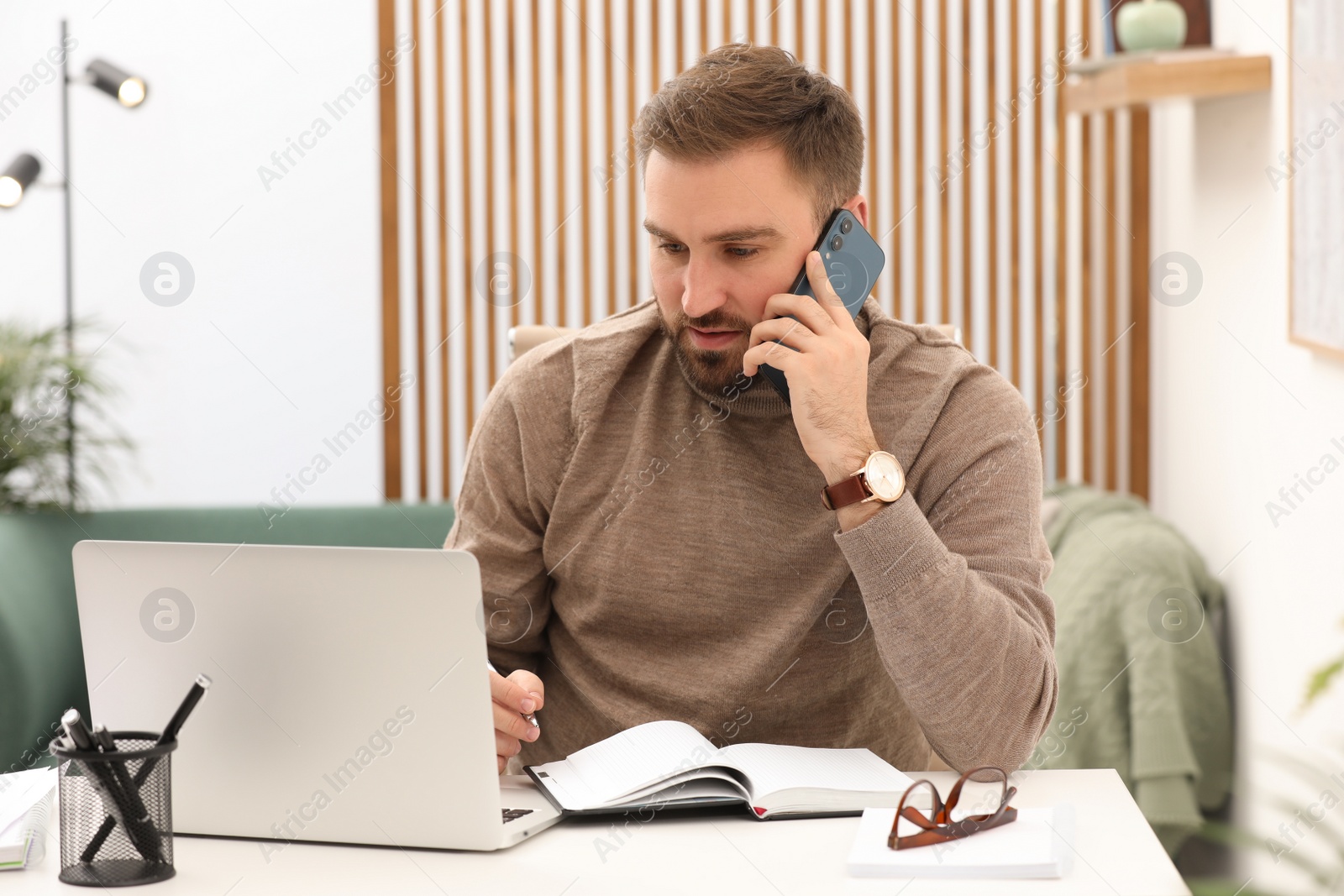 Photo of Young man talking on phone while working with laptop at home