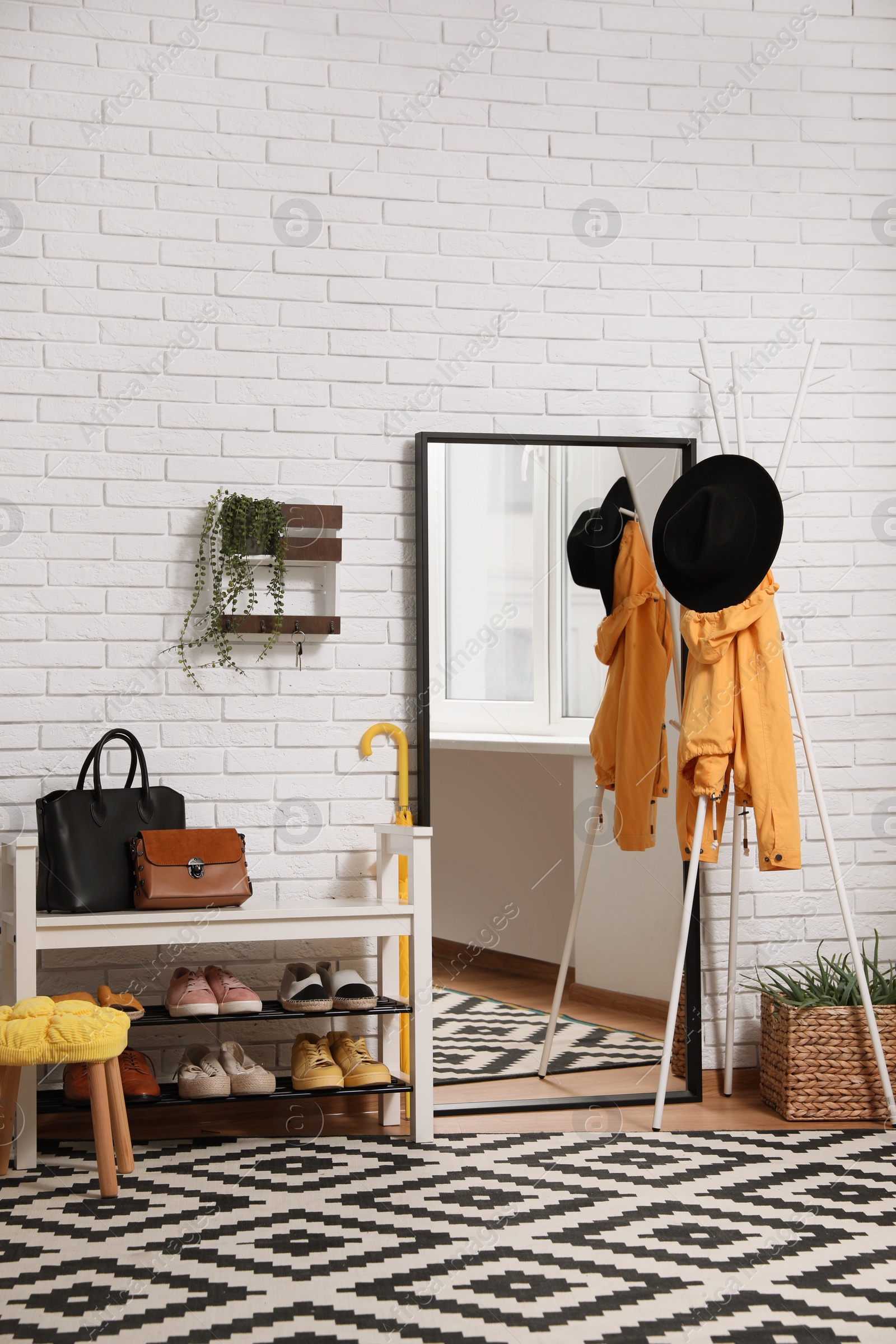 Photo of Stylish hallway interior with coat rack, shoe storage bench and mirror near white brick wall