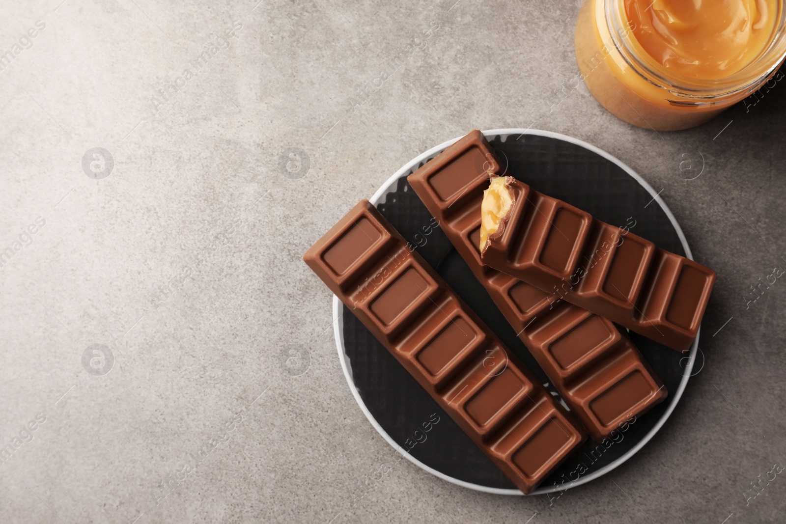 Photo of Tasty chocolate bars and jar of caramel on grey table, flat lay. Space for text