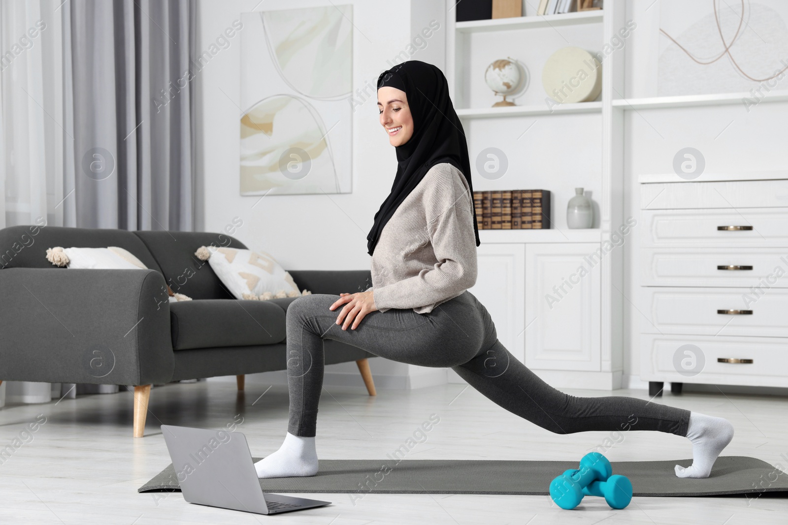 Photo of Muslim woman in hijab stretching near laptop on fitness mat at home