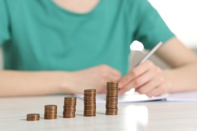 Financial savings. Woman making notes at white wooden table, focus on stacked coins