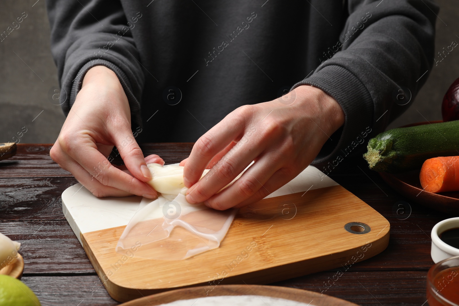 Photo of Making delicious spring rolls. Woman wrapping fresh cabbage into rice paper at wooden table, closeup