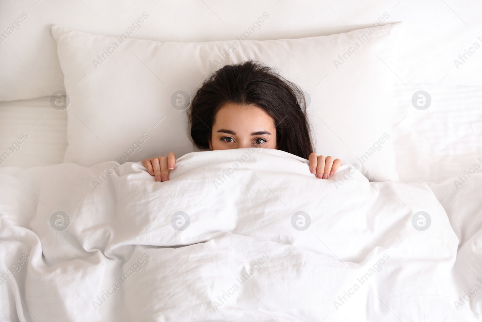 Photo of Young woman hiding under warm white blanket in bed, top view