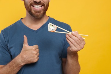 Photo of Happy man holding sushi roll with chopsticks and showing thumbs up on orange background, closeup