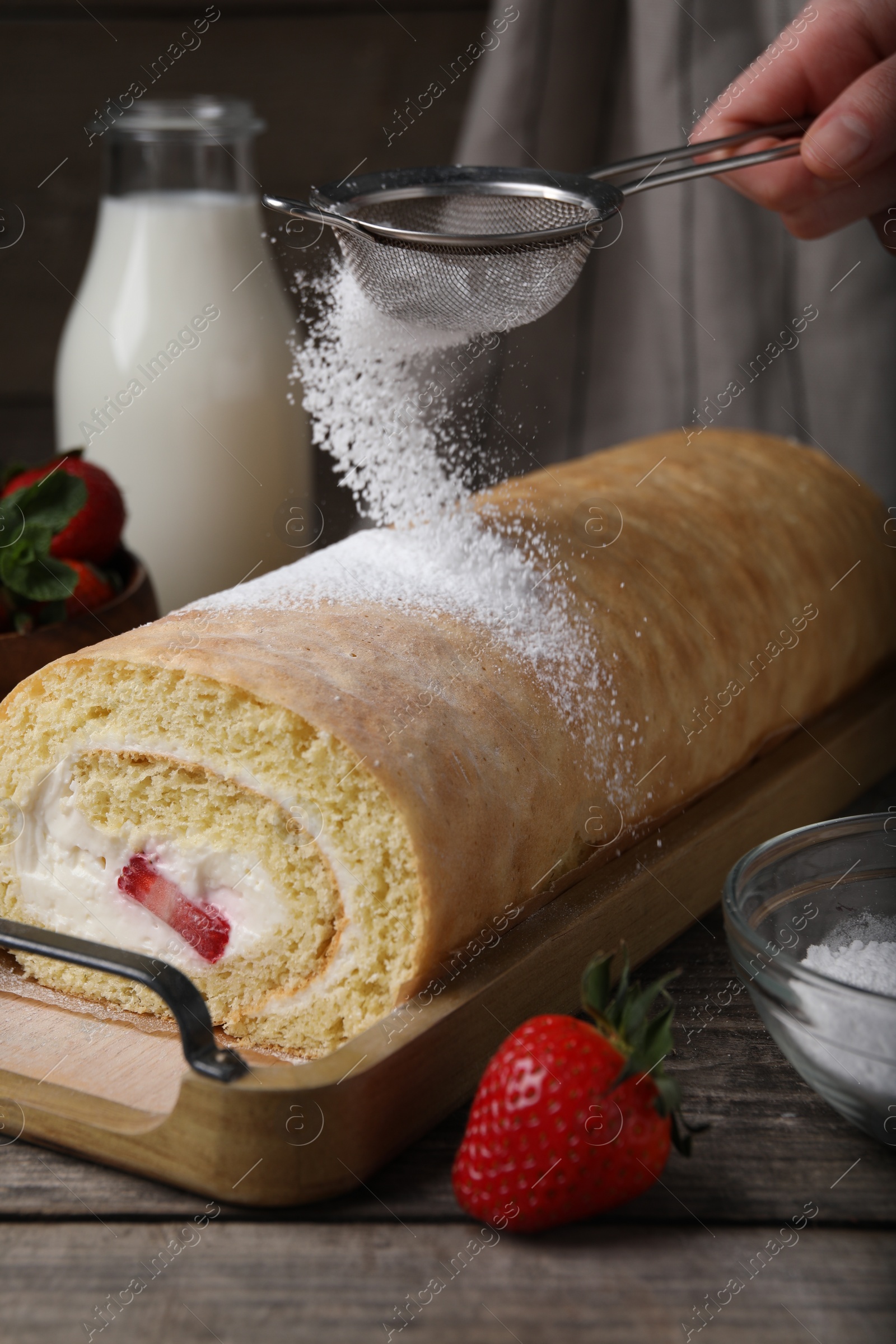 Photo of Woman pouring powdered sugar onto delicious sponge cake roll with strawberries and cream at wooden table, closeup