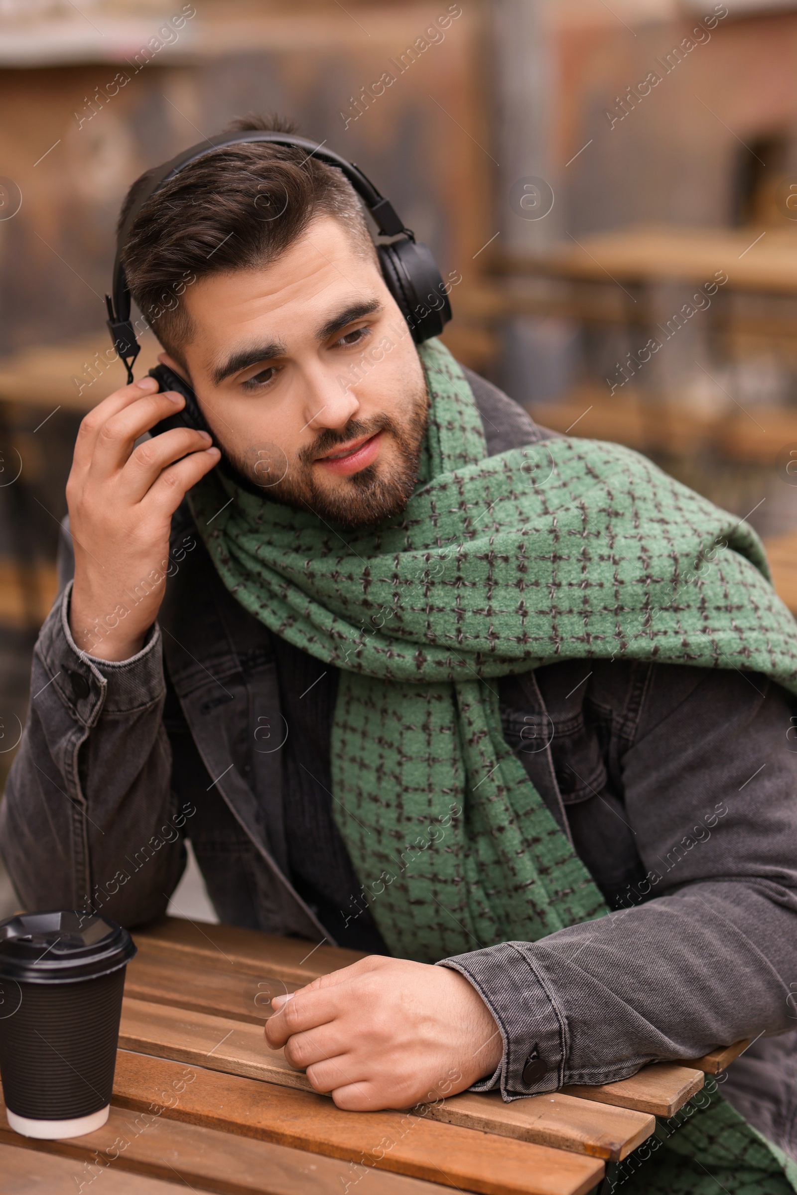 Photo of Handsome man in warm scarf with paper cup listening to music in outdoor cafe