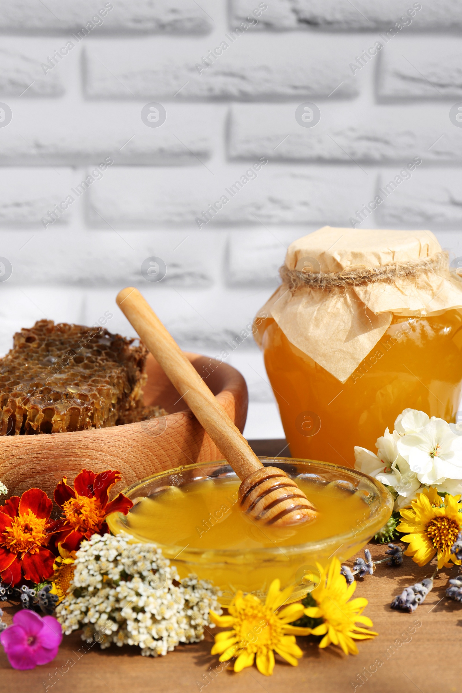 Photo of Delicious honey, combs and different flowers on wooden table near white brick wall