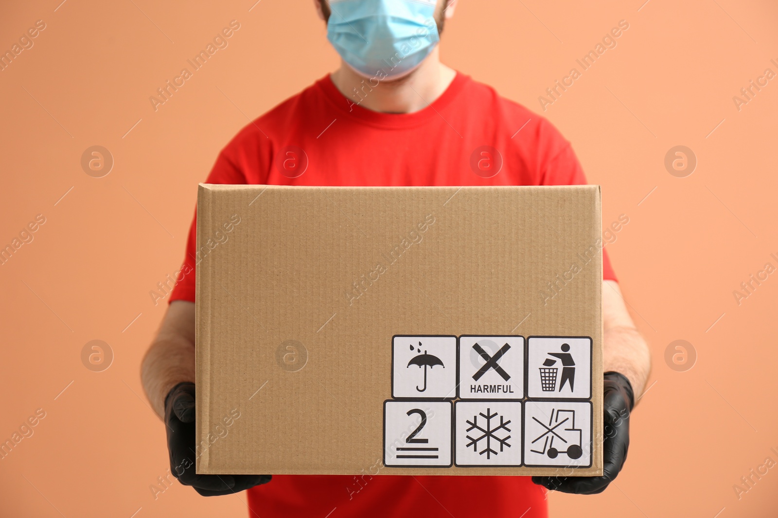 Photo of Courier holding cardboard box with different packaging symbols on orange background, closeup. Parcel delivery
