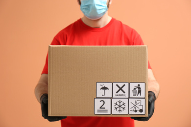 Courier holding cardboard box with different packaging symbols on orange background, closeup. Parcel delivery