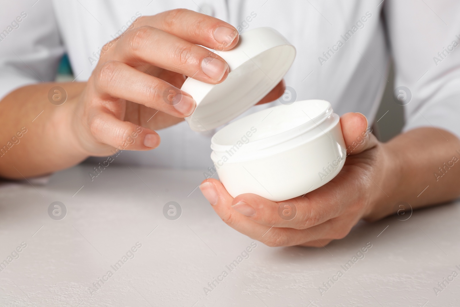 Photo of Dermatologist holding jar of cream at white table, closeup. Developing cosmetic product