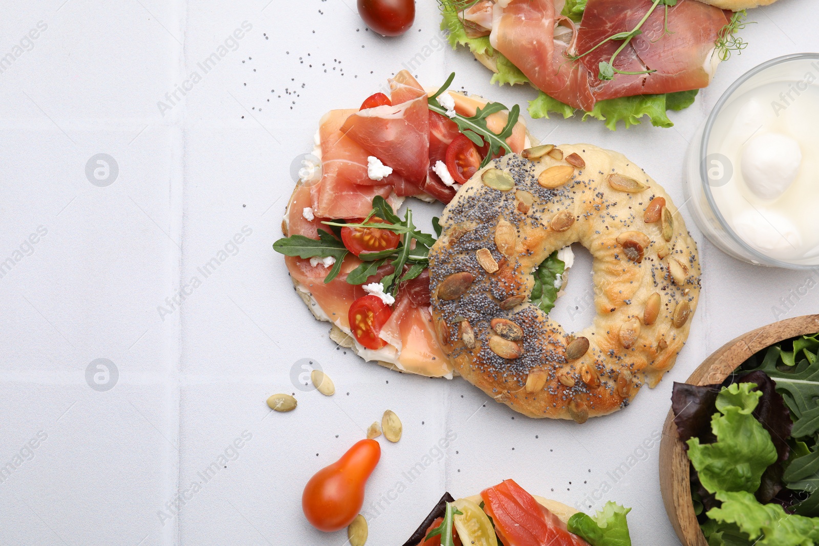 Photo of Tasty bagel with cured ham, cheese, tomatoes and arugula on white tiled table, top view. Space for text